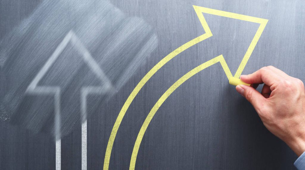 A arrow drawn in yellow chalk on a chalk board turning away from a previously drawn and erased white arrow.