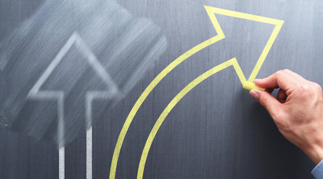 A arrow drawn in yellow chalk on a chalk board turning away from a previously drawn and erased white arrow.