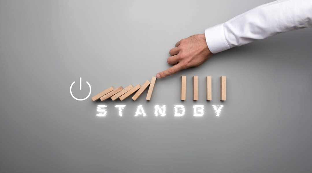 "STANDBY" written under a person interrupting the flow of wooden domino bricks by holding up the falling one.