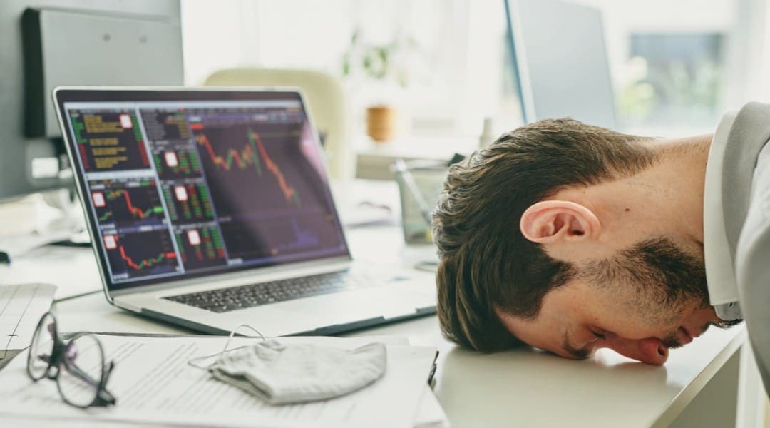 a person resting his head at his desk in front of his laptop displaying various graphs.