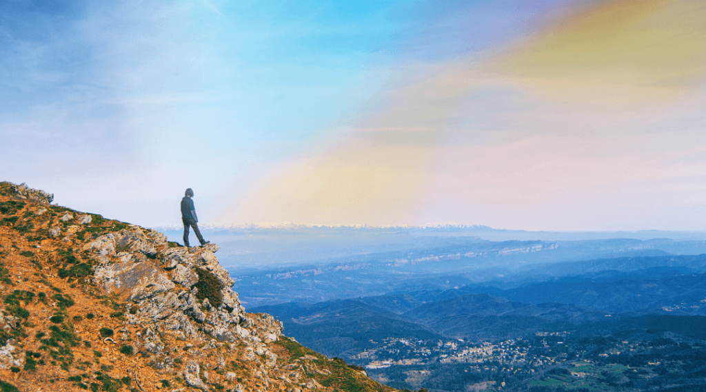 A person on a mountain cliff looking at a view of smaller green mountains and an orange and blue skyline