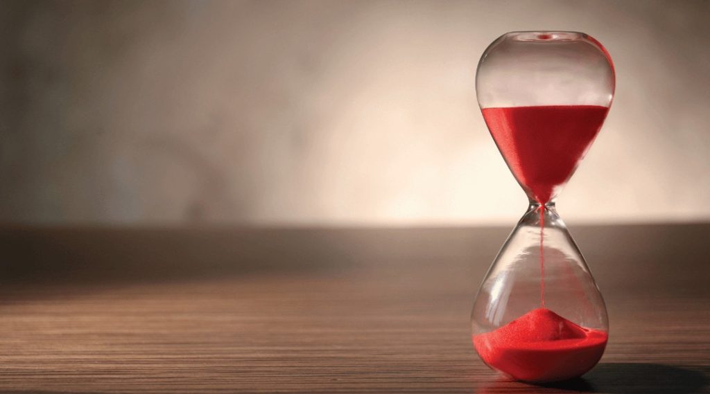 a close up of a hour glass time with red sand half done sitting on a wooden table.