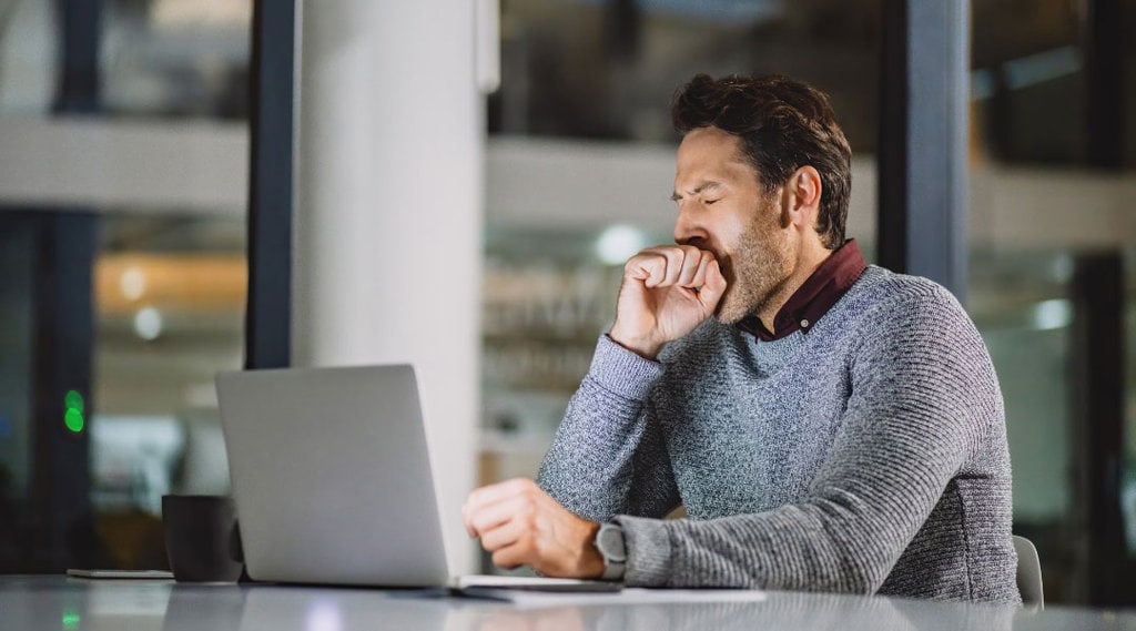 A person looking at his computer while holding his hand over his mouth and yawning