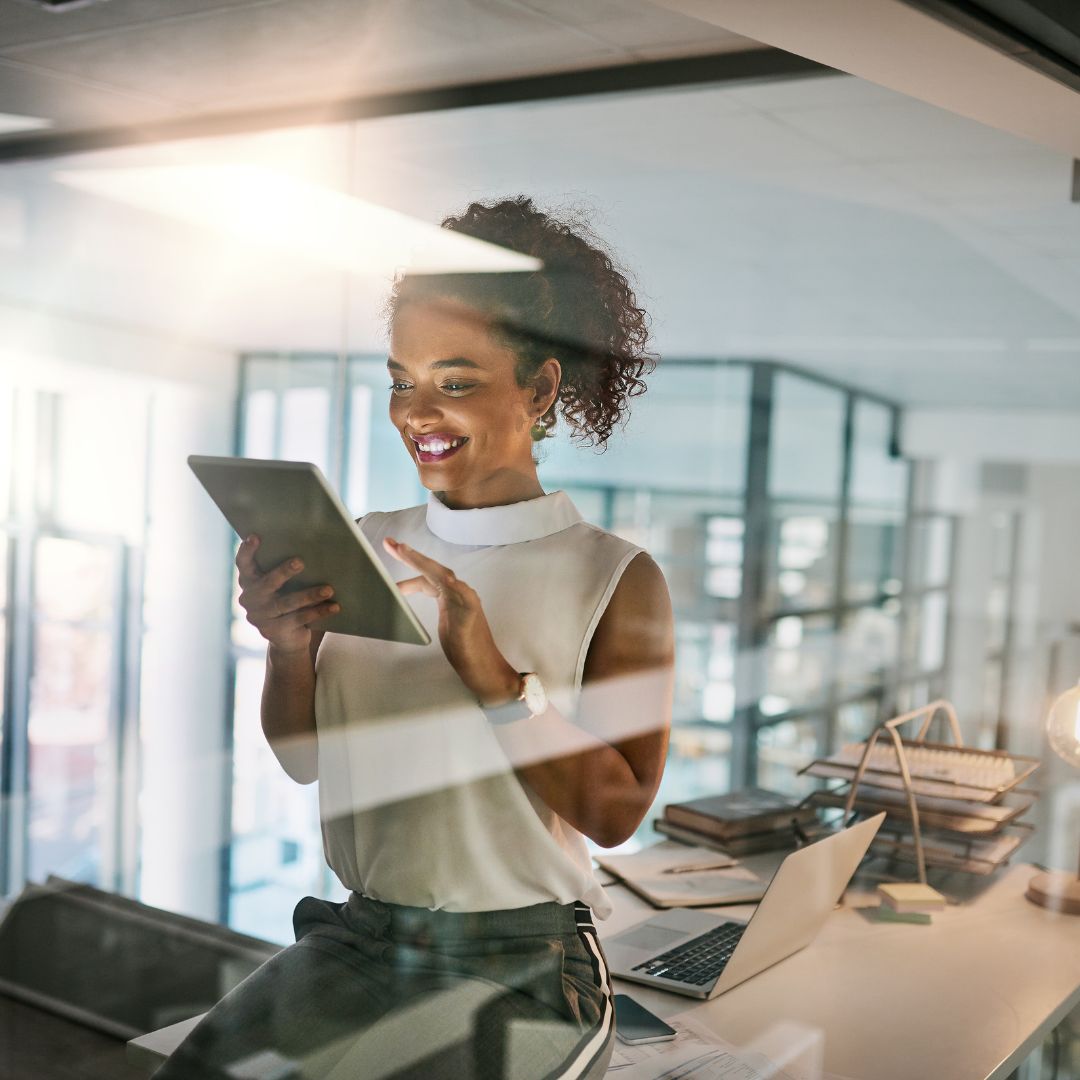 professional individual sitting on desk in office, smiling at her lit up tablet screen