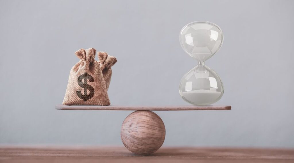 A burlap money bag with a dollar sign and a glass hourglass balancing on a wooden seesaw.