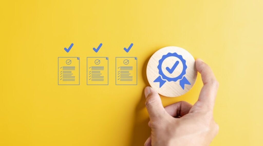 Hand holding a wooden badge with a blue checkmark, next to illustrated checklists and checkmarks on a yellow background, representing quality assurance or compliance.
