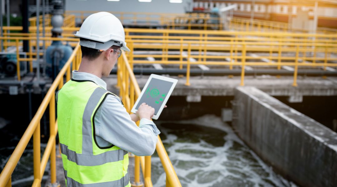 Engineer in safety vest and helmet using a digital tablet while inspecting a water treatment facility with yellow railings and flowing water.